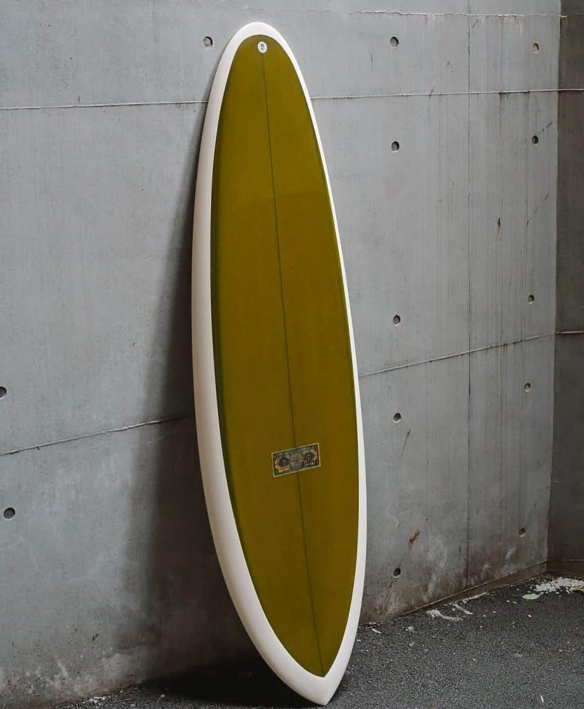 shapers-club- Une planche de surf Jeff McCallum jaune-vert avec un bord blanc est posée à la verticale contre un mur de béton gris. La surface sous la planche de surf est sombre et légèrement texturée. -surf