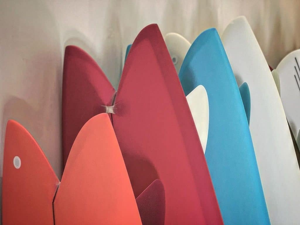 shapers-club- Gros plan sur plusieurs planches de surf colorées de Jeff McCallum, debout en rangée, mettant en valeur leurs ailerons pointus et leurs bords dans des tons de rouge, d'orange, de rose, de bleu et de blanc. -surf