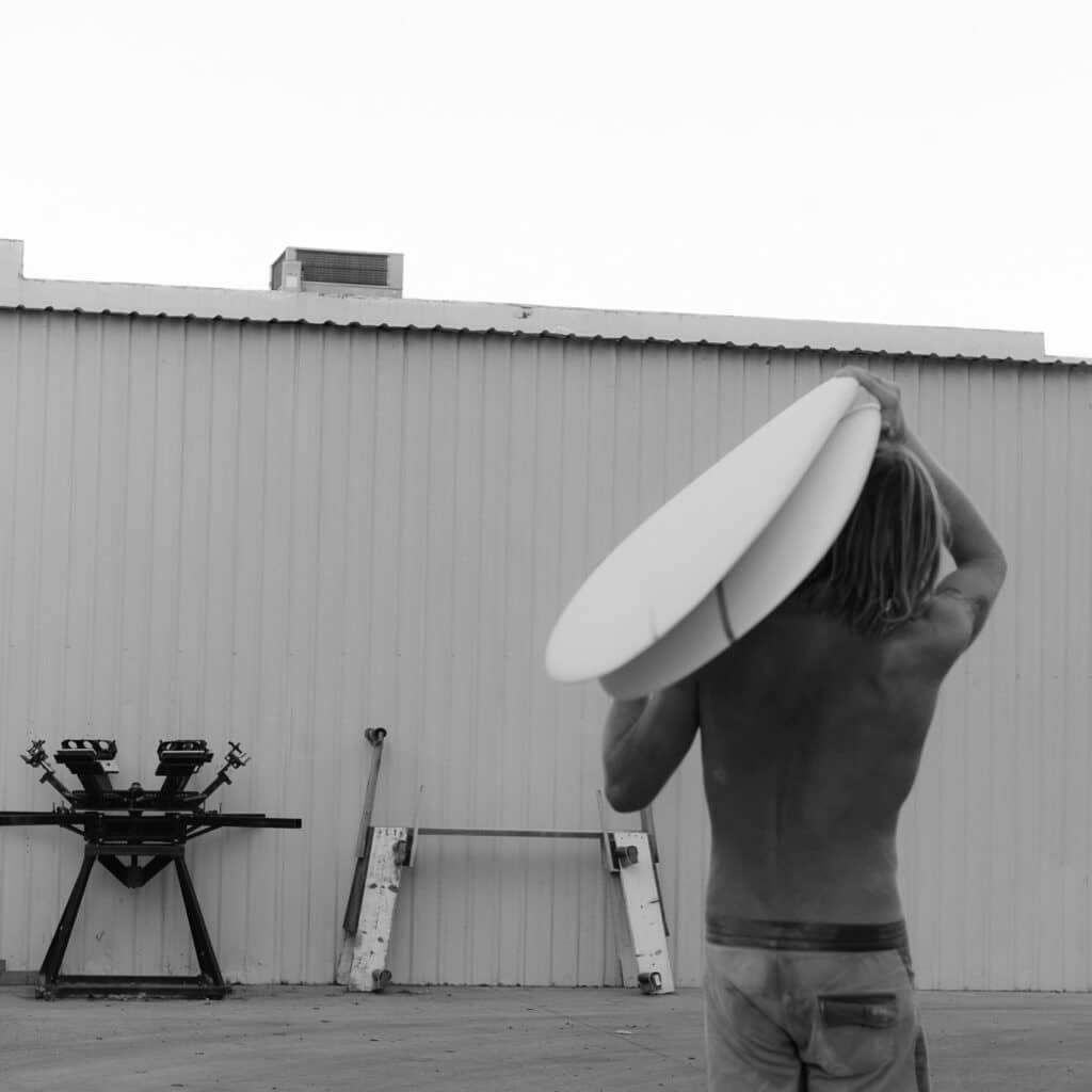 shapers-club- Une personne torse nu aux cheveux longs, peut-être Kai Ellice-Flint, porte une planche de surf sur son épaule en direction d'un bâtiment en tôle ondulée du Shapers Club. Sur cette image en noir et blanc, deux chevalets et des planches en bois sont appuyés contre le mur. -surf