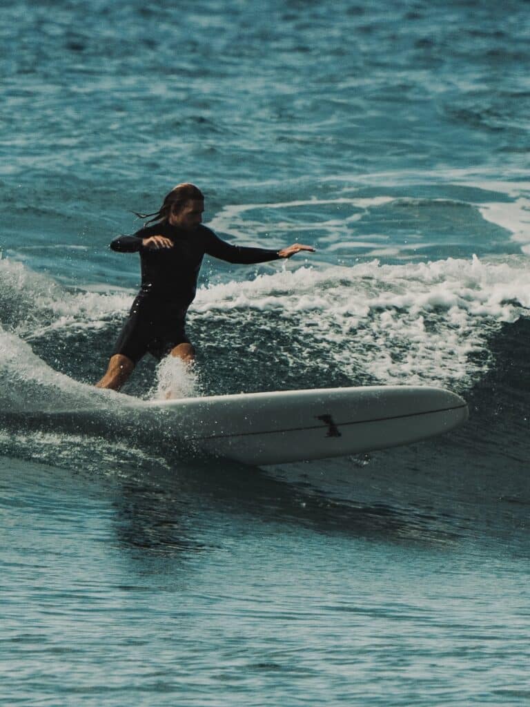 shapers-club- Une personne vêtue d'une combinaison noire chevauche une planche de surf blanche sur une petite vague de l'océan, en équilibre avec les bras tendus. L'eau est bleue avec de l'écume blanche près de la planche. Style classique inspiré du Shapers Club et de sa résidence de shape. -surf
